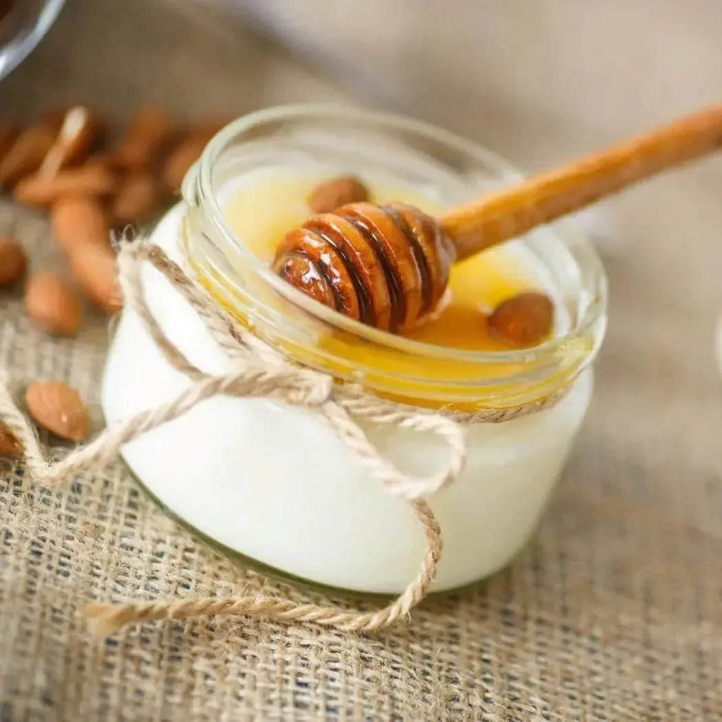 A glass jar of creamy vanilla and honey blend with a honey dipper, surrounded by almonds.