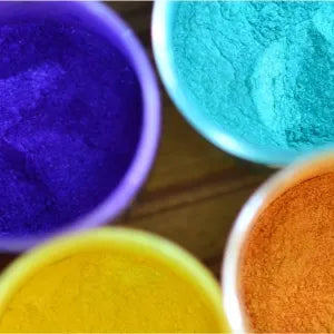 Vibrant, finely textured pigment powders in purple, teal, yellow, and orange hues are contained within individual plastic cups.