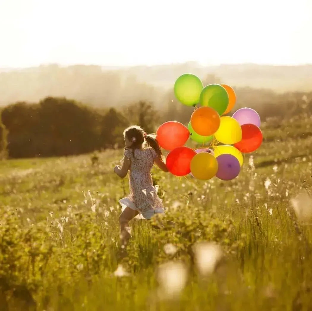 A girl runs joyfully in a field, holding a colorful bouquet of balloons against a sunny backdrop.