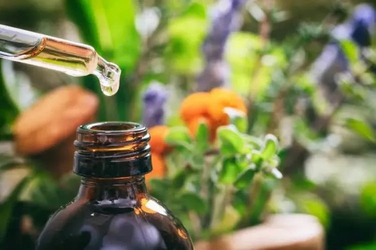 Glass dropper releasing essential oil into a brown bottle, surrounded by greenery and colorful flowers.