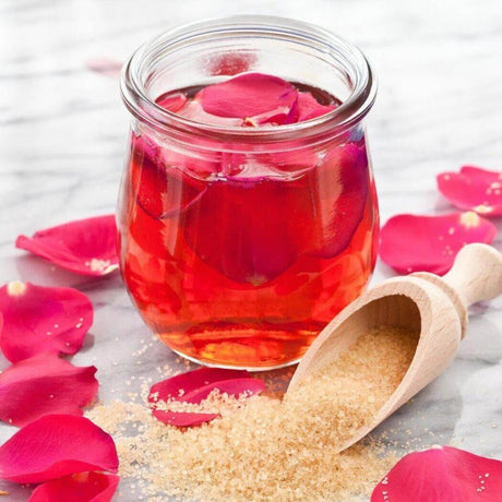 Jar of handmade rose petal infusion with scattered rose petals and brown sugar scoop on marble surface - Nurture Handmade.