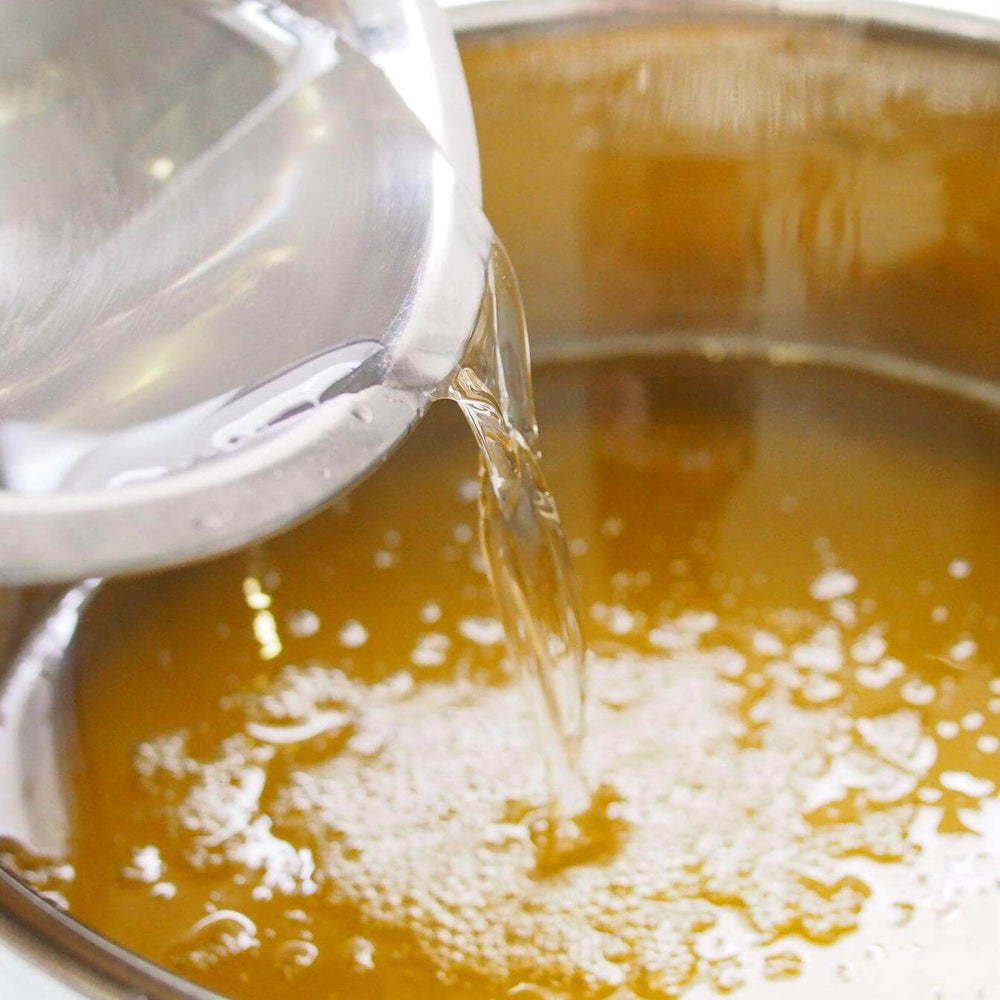 Clear glass pitcher pouring water into a pot of golden liquid.