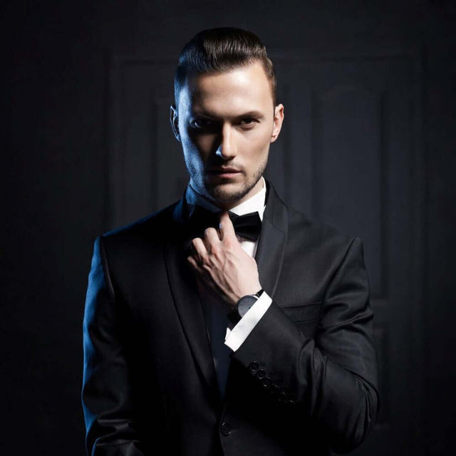 Elegant man in a black tuxedo, adjusting his bow tie, standing with a serious expression in a dimly-lit room.