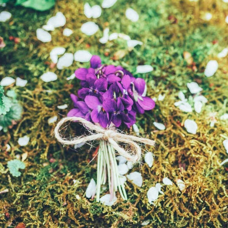 A small bouquet of purple flowers with a twine wrap, resting on a lush green mossy ground, surrounded by scattered white petals. Nurture Handmade