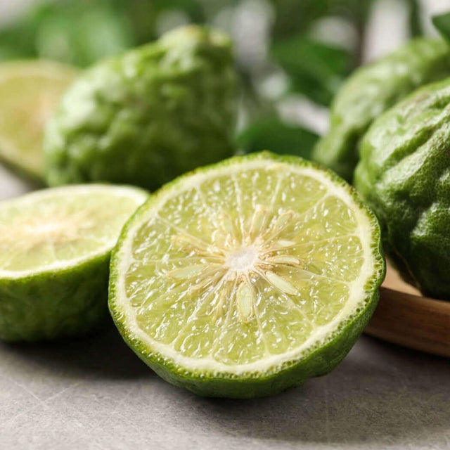 Fresh green kaffir lime halved with whole limes in the background, showcasing textured rind and juicy interior. Nurture Handmade.