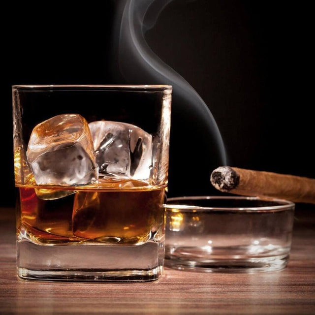 Glass of whiskey with ice beside a smoking cigar in an ashtray on a wooden table