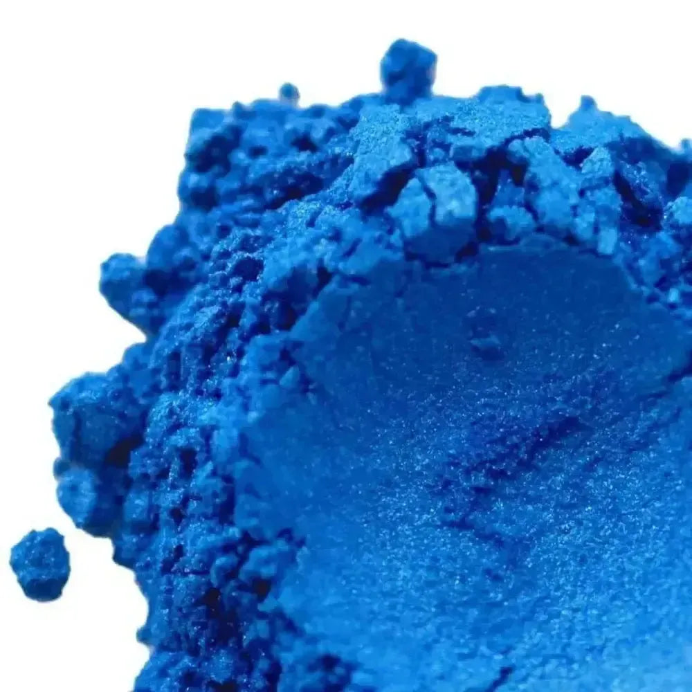 A close-up of Brilliant Blue Mica pigment powder reveals its vibrant color and textured, crumbly surface against a white background.