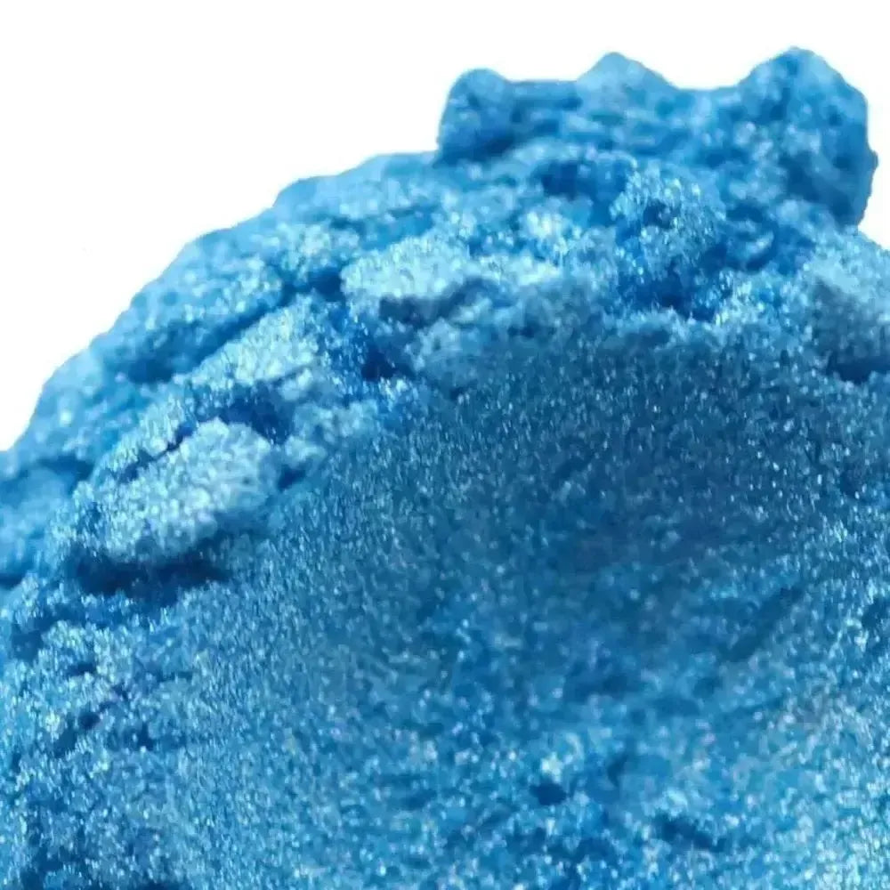 A close-up of shimmery Caribbean Mica eyeshadow shows its textured, powdery surface with sparkling, reflective particles and a vibrant tropical blue tone.