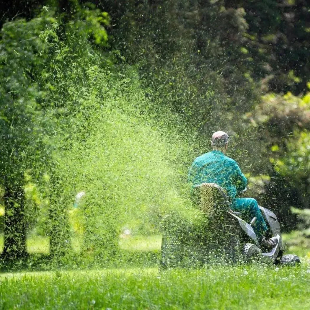 A man in a teal outfit mows grass, creating a cloud of fresh-cut greenery on a sunny day.