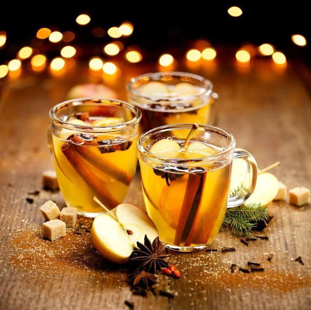 Three glasses of spiced apple cider with cinnamon sticks and star anise, surrounded by apple slices and sugar cubes, on a festive wooden table.