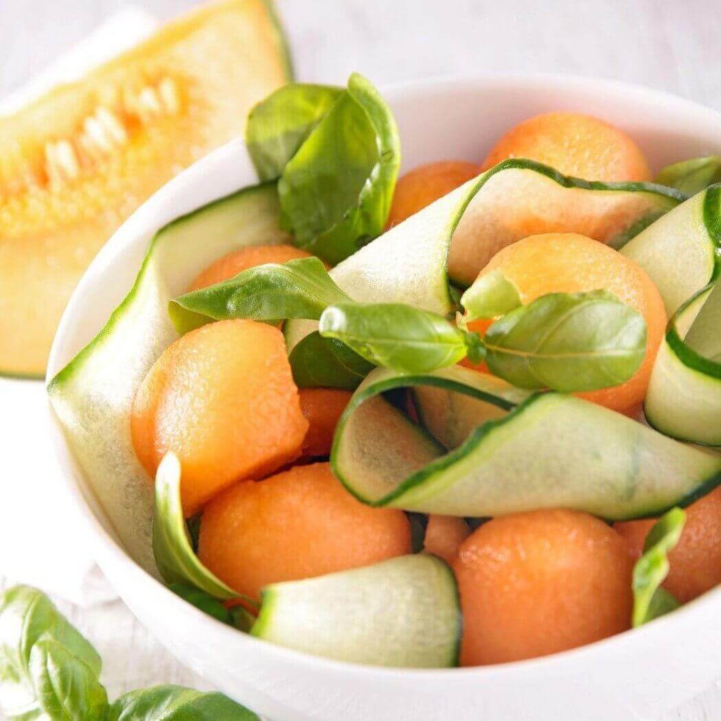 Fresh cantaloupe balls with cucumber ribbons and basil leaves in a white bowl, showcasing a healthy, handmade summer salad.