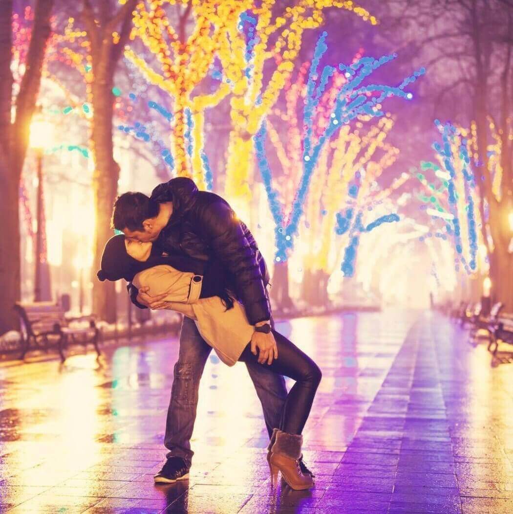 Couple sharing a romantic kiss under vibrant, colorful holiday lights in a serene, night-time park setting.