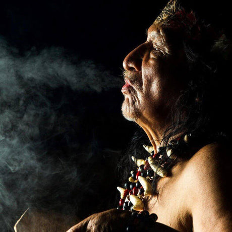 A person with traditional jewelry and headwear blowing smoke into the dark, highlighting cultural practices and reverence.