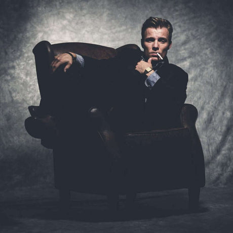 Man sitting in an armchair with a serious expression in a dark, moody setting.