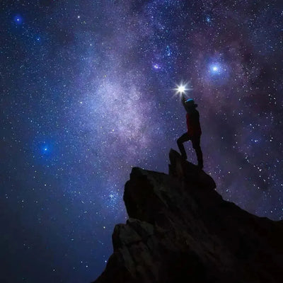 Silhouetted hiker on rocky peak holding light beneath starry Milky Way