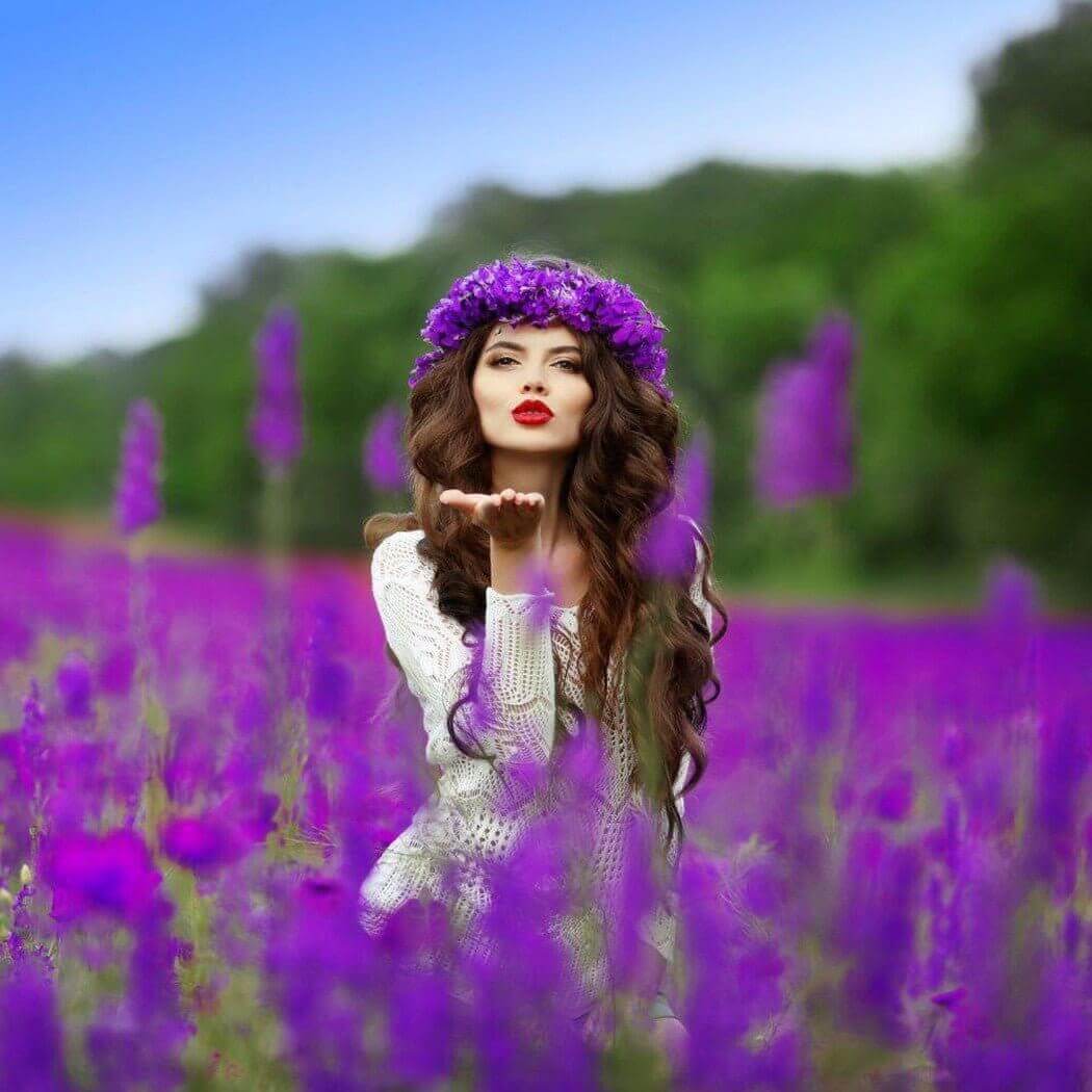 Woman in white dress and purple flower crown blowing a kiss in a field of purple flowers, showcasing a serene and nature-inspired scene.