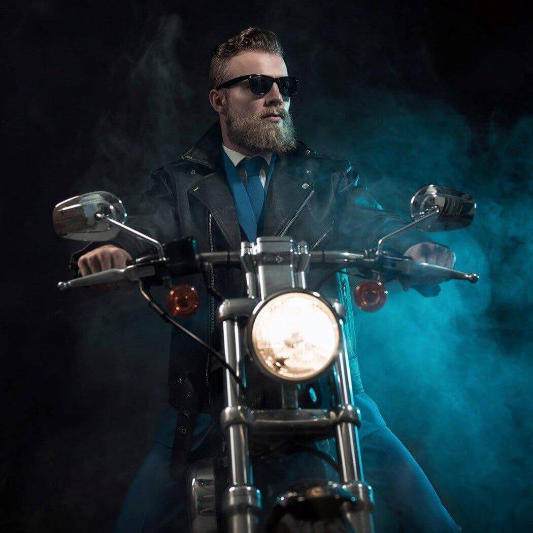 Bearded man in sunglasses and leather jacket sitting on a motorcycle surrounded by blue smoke.