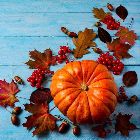 Bright orange pumpkin on blue wooden background surrounded by autumn leaves, acorns, and red berries, reflecting Nurture Handmade aesthetic.