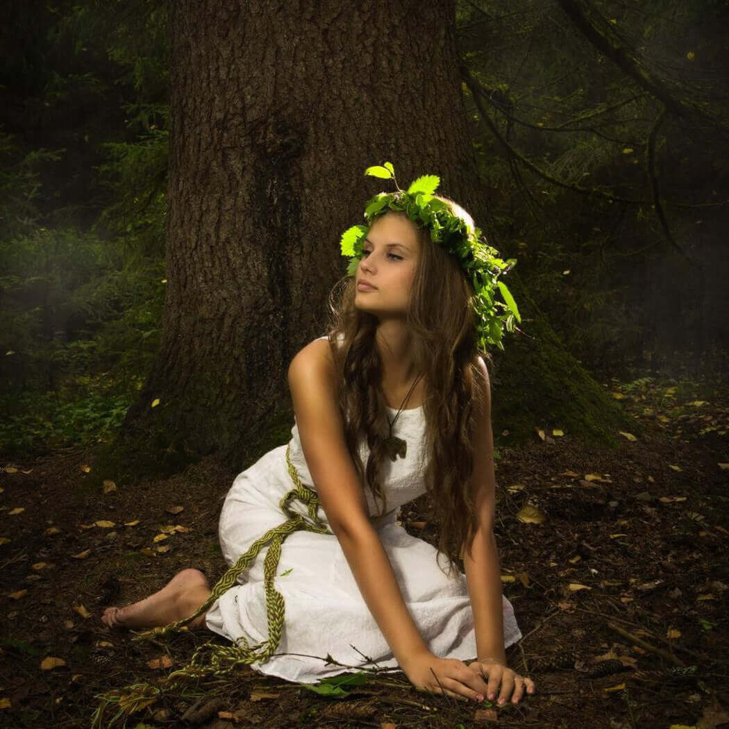 Woman in white dress and green leaf crown sitting by a tree in the forest, representing Nurture Handmade natural and organic lifestyle.