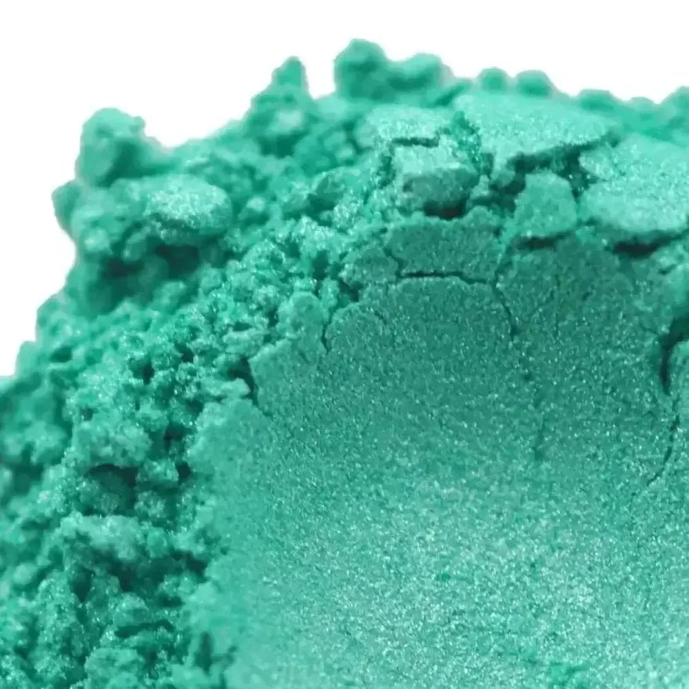 A close-up of Sea Green Blue Mica eyeshadow shows its bright teal green shade, shimmery metallic finish, powdered texture, and a small indentation in the center.