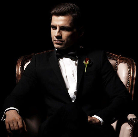 Man in a tuxedo sitting on a vintage chair in a dark setting with a red rose pinned to his lapel.