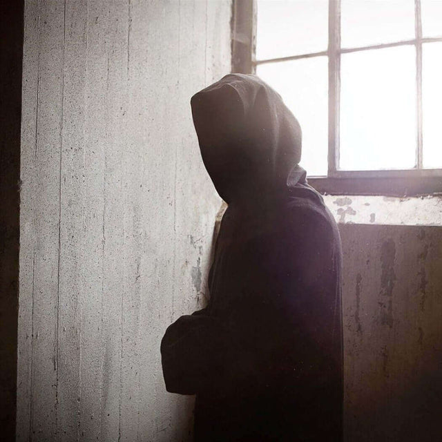 Hooded figure standing alone by a window in a dimly lit room, facing away from the camera.