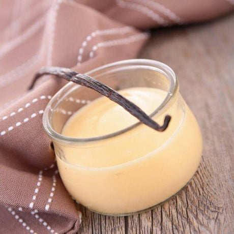 Handmade vanilla pudding in a glass jar with vanilla bean garnish, placed on a rustic wooden table with a brown dotted nappe, Nurture Handmade.