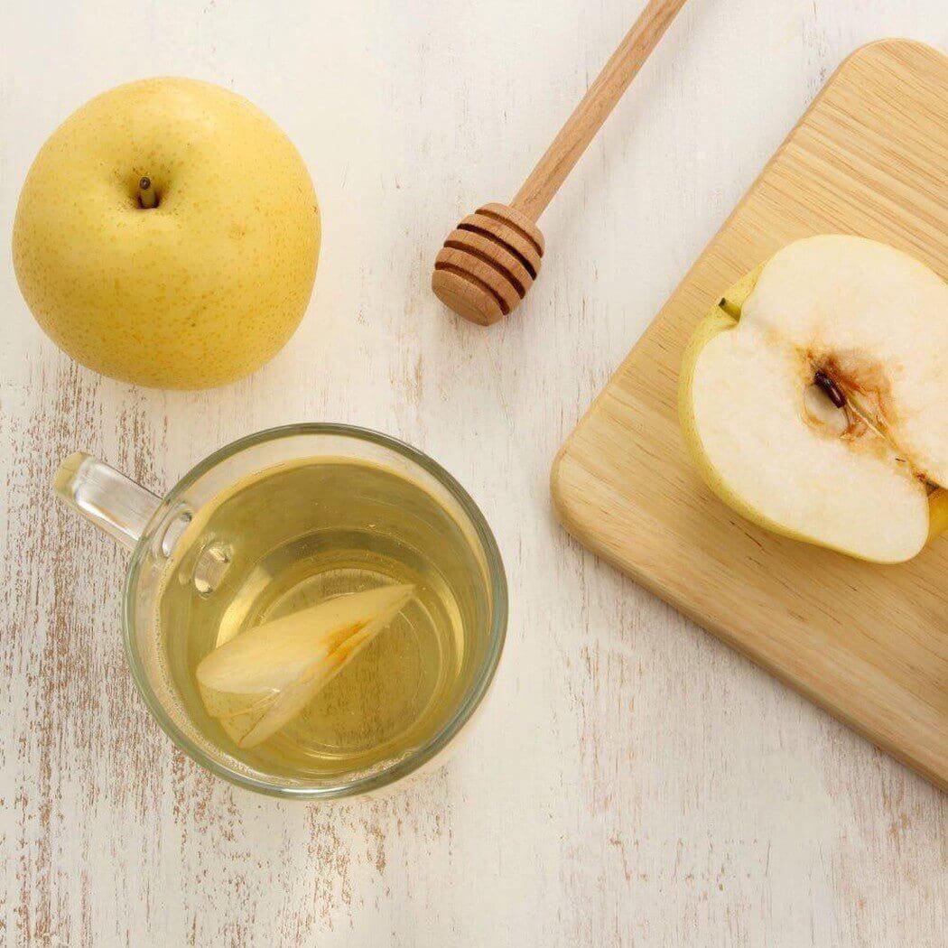 A cup of pear tea with a fresh whole pear, a sliced pear on a cutting board, and a honey dipper on a wooden table. Nurture Handmade ingredients.